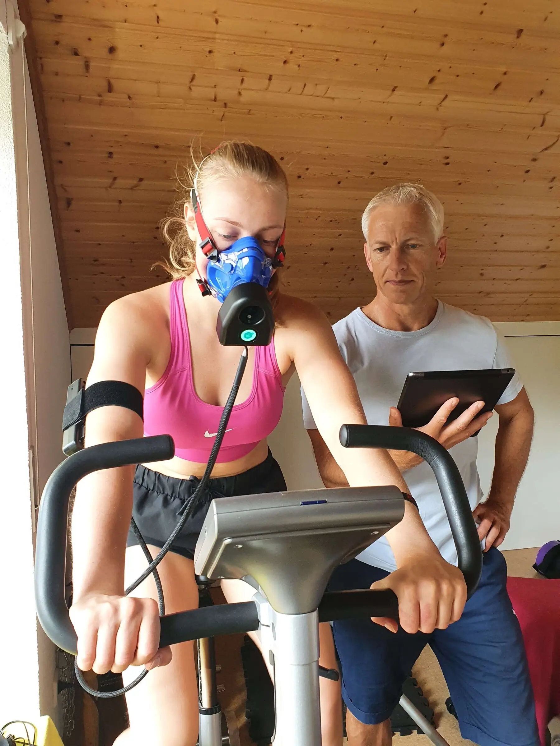 Sportlerin fährt mit Atemmaske auf dem Ergometer, Uwe Schapeit verfolgt die Messwerte auf dem Tablet