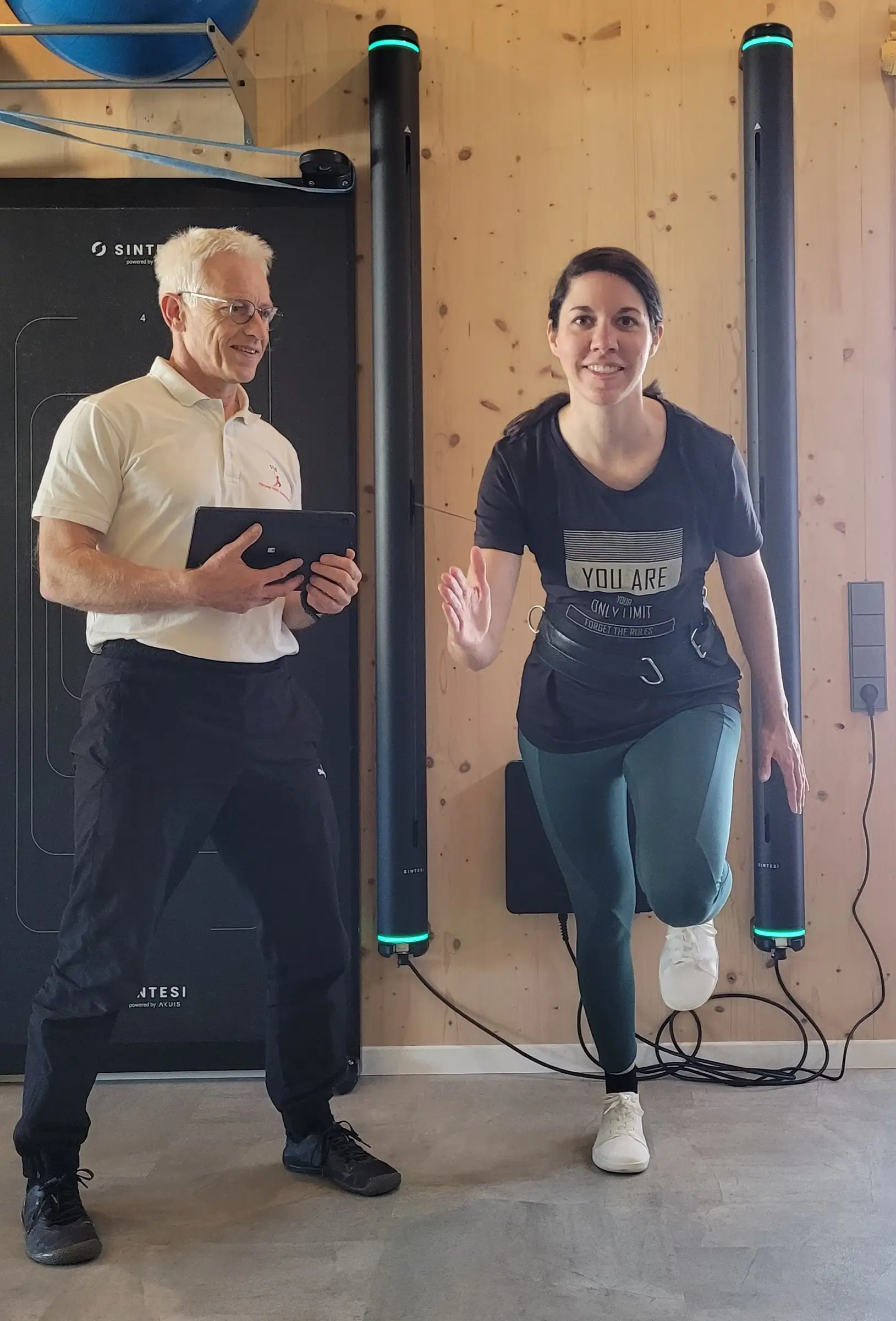 Uwe Schapeit mit Tablet neben einer Kundin, die am Sintesi-Gerät auf einem Bein eine Laufbewegung übt