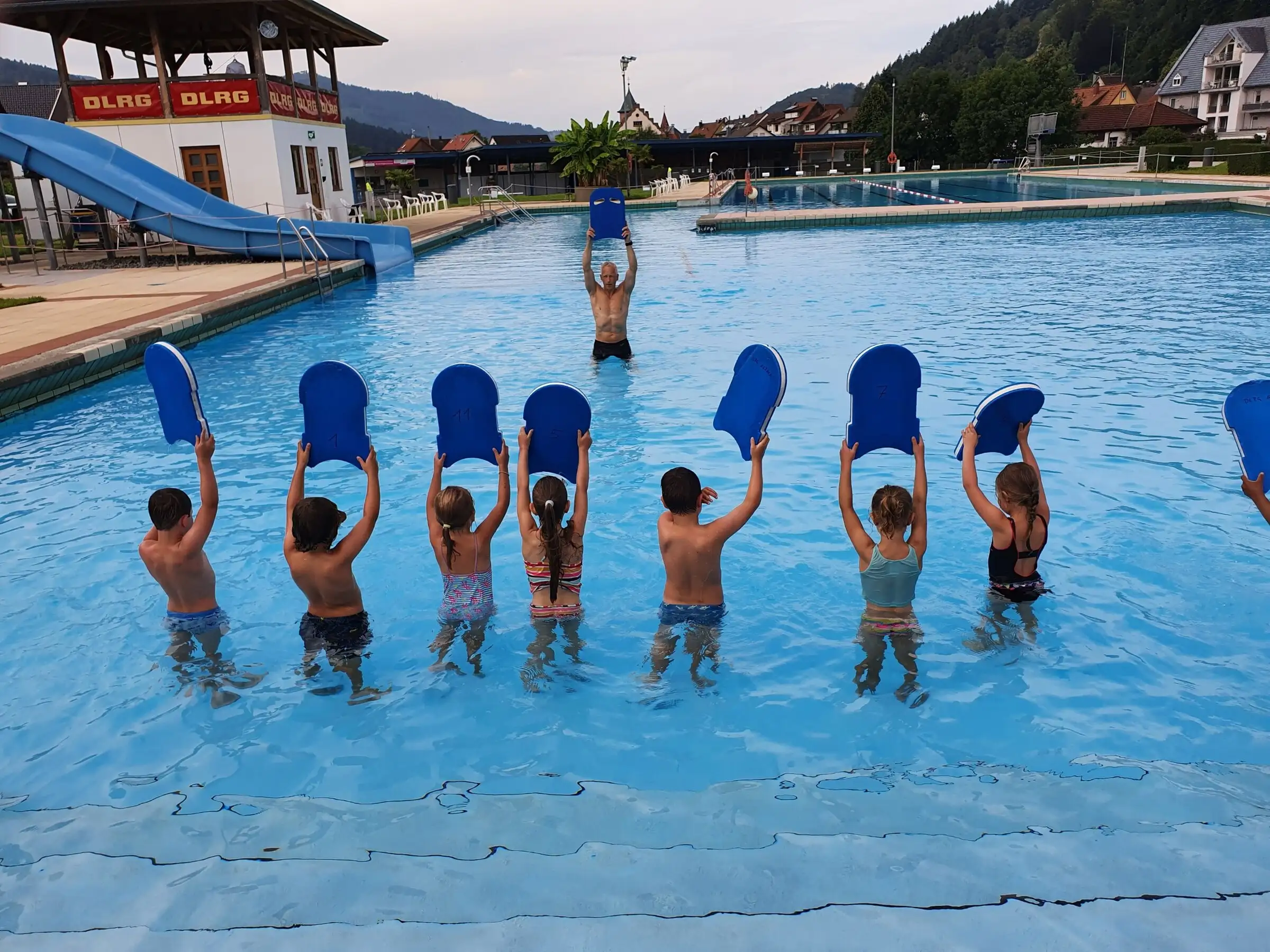 Kinder im Schwimmkurs halten im Freibad Schwimmbretter in die Höhe, von hinten fotografiert