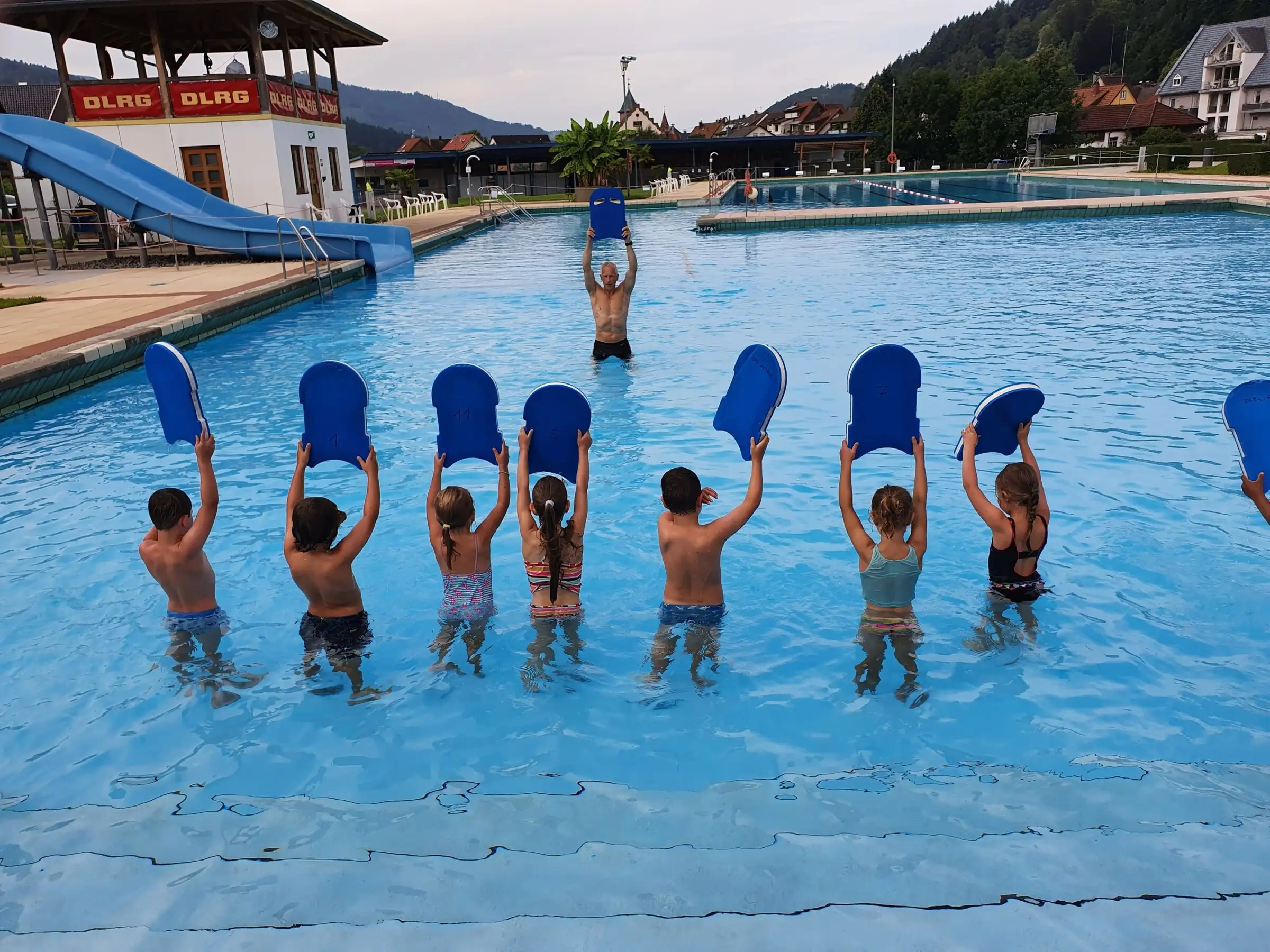 Kinder im Schwimmkurs halten im Freibad Schwimmbretter in die Höhe, von hinten fotografiert