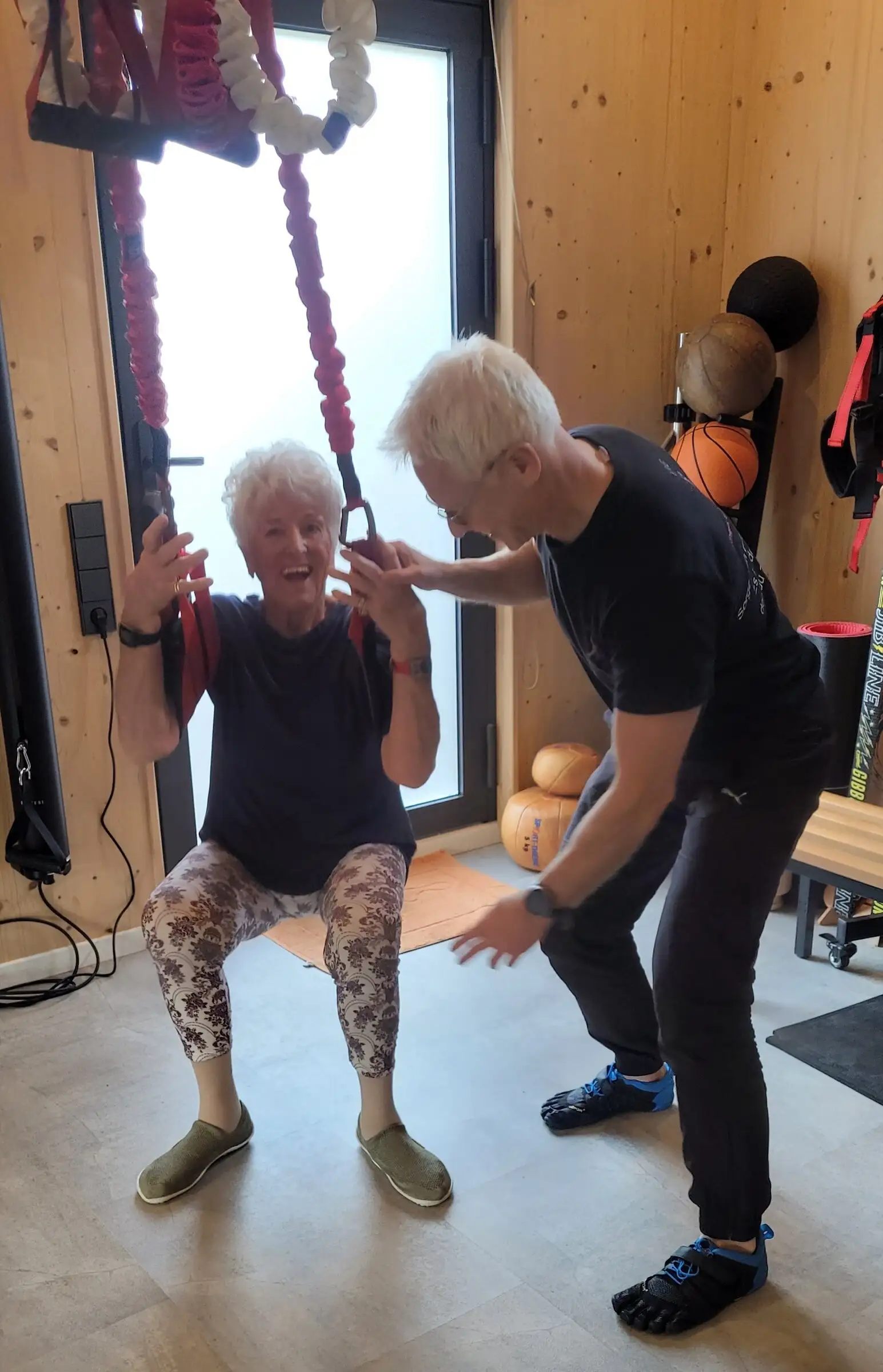 Lachende Seniorin in der Hocke am 4D PRO Schlingentrainer, Uwe Schapeit sichert sie