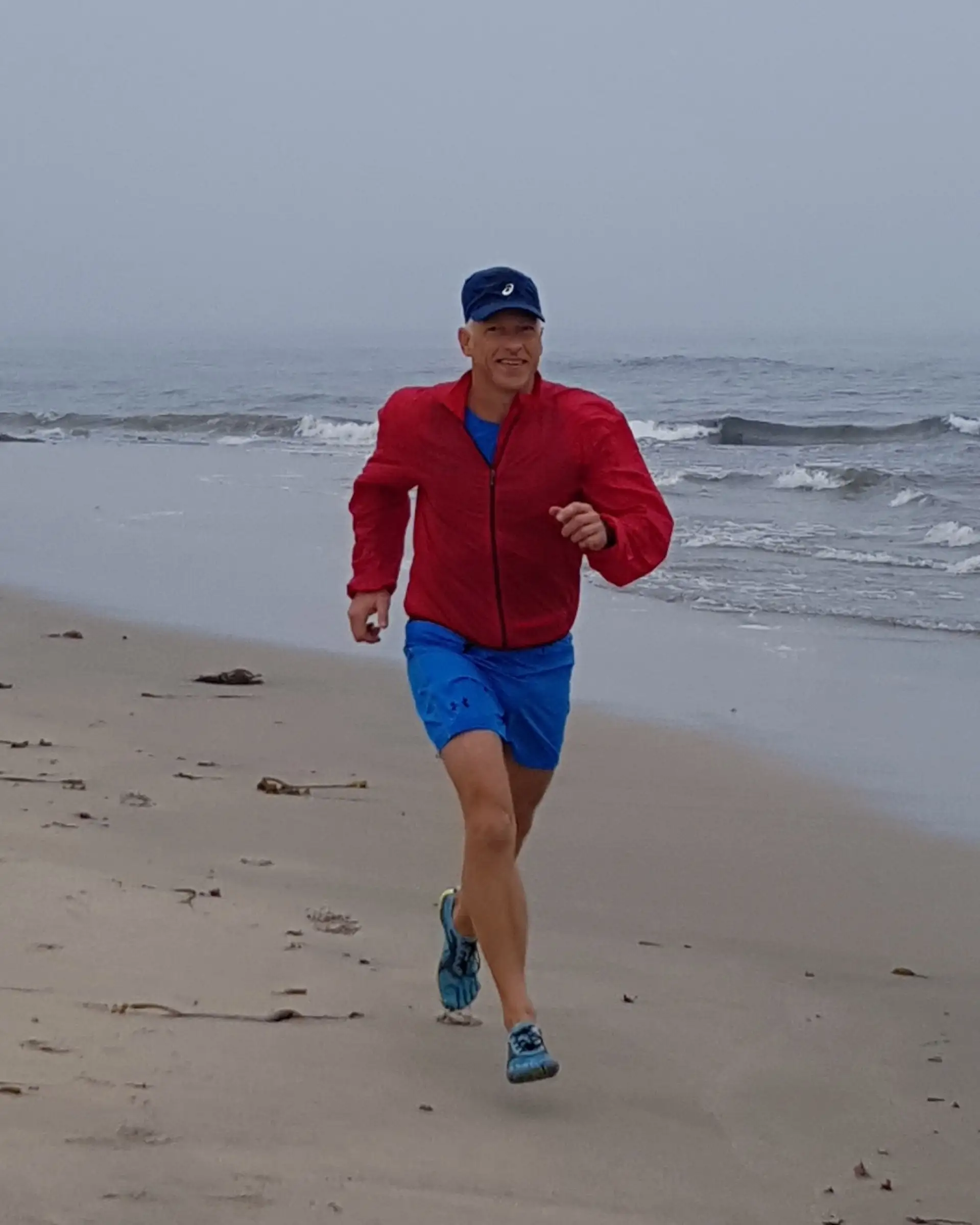 Uwe Schapeit läuft in roter Jacke barfuß am Strand entlang